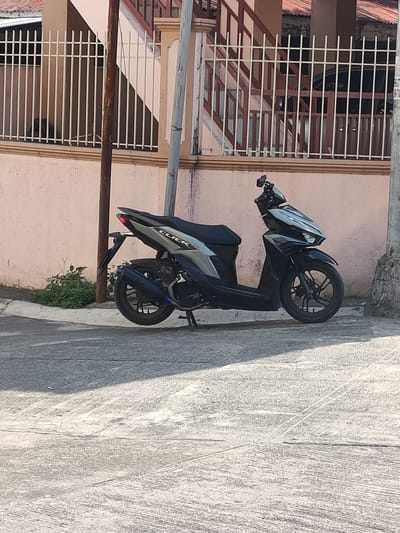 A black and silver Honda Click scooter is parked on a concrete road next to a beige wall and a white metal fence. The scooter is angled to the right, with its rear tire closest to the viewer. The scooter has a dark blue exhaust pipe with a chrome accent. Behind the scooter, there is a patch of green bushes. A utility pole stands to the right of the scooter. In the background, a building with a reddish-brown roof and a brown and white railing is visible. The scene appears to be in a residential area in San Pascual, Philippines, as indicated by the scooter model and the architectural style.