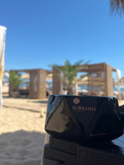 This is a close-up, eye-level, outdoor shot of a black ashtray with the "SOBRANIE LONDON" logo in gold on a textured surface, likely part of a chair or table. The ashtray is in focus and angled slightly towards the viewer, with a curved indentation visible on its surface, suggesting it's designed for holding cigarettes. Behind the ashtray, the scene is blurred and out of focus, depicting a beach environment. Several open-air wooden structures with thatched roofs, possibly beach cabanas or bar areas, are visible in the mid-ground, along with palm trees. The background is dominated by a clear, bright blue sky. In the far background, faint figures of people and the sea can be discerned. The overall impression is a sunny day at a beach resort. The text on the ashtray, "Пушенето убива – отказвате се от себе си", is a Bulgarian warning which translates to "Smoking kills - you give up on yourself." The setting is consistent with a beach resort in Dyuni, Bulgaria, known for its coastline and beach amenities.