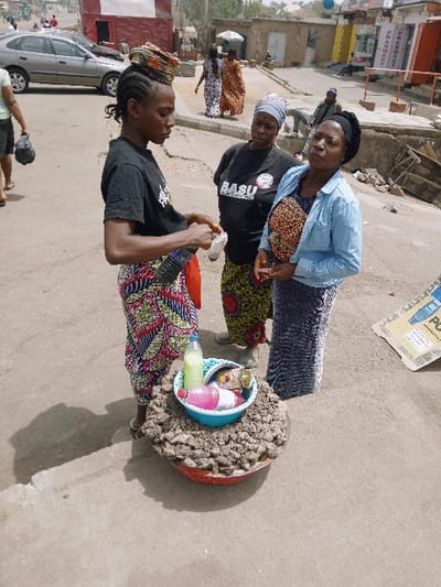 This image captures an outdoor street scene in Kadoma, Zimbabwe, under bright daylight. In the foreground, a young woman with braided hair, wearing a black t-shirt, a patterned skirt, and a colorful headwrap, holds a clear plastic bottle in her left hand and a small white packet in her right, appearing to open or prepare an item. On the ground in front of her is a large, shallow, red-rimmed basin filled with numerous small, light brown, irregular objects. Resting within this basin is a smaller blue bowl containing several plastic bottles of liquids (one light yellow, one pink, and one clear with brown contents) and a small metallic container.

To the right of the foreground woman, two other adult women are present. The woman closer to the foreground wears a black t-shirt with "BASU" and a logo, a patterned skirt, and a light blue headwrap; she is looking towards the foreground woman. The woman furthest to the right wears a light blue denim jacket over a leopard-print top and a dark patterned skirt, with a dark headwrap; she holds a small pink object in her right hand and is looking slightly away.

The ground surface is an uneven, light-colored material, possibly dirt or unpaved concrete. In the background, a silver sedan is parked on the left side of a street. Beyond this, several single-story buildings are visible, some with signs, including a partially legible "PHARMACY" sign. Other individuals are present in the deeper background, including two women walking on a raised platform and a man seated on a ledge. Distinct shadows are cast on the ground, indicating direct sunlight.
