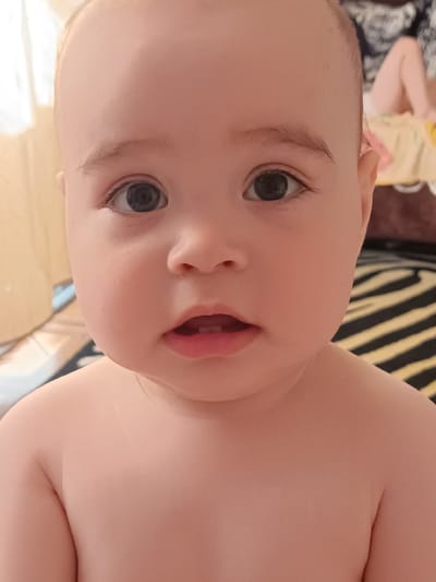 The image is a close-up portrait of a baby's face, with the shoulders and upper chest visible. The baby has large, dark eyes and small lips with two visible teeth. The background is blurred, showing some striped fabric and a hint of a person's legs. The lighting is soft and natural. There is no discernible indication of the location being in the city of Посад, Russia.