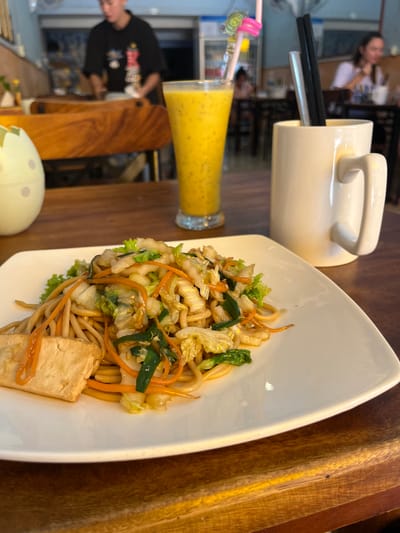A forensic examination of the image reveals a dining scene inside a restaurant located in Khan Daun Penh, Cambodia. In the foreground, a white rectangular plate holds a serving of stir-fried noodles, prepared with thick, light brown noodles, green cabbage, sliced orange carrots, and green onions. A rectangular piece of fried tofu rests on the left side of the plate.
To the right of the plate, on a dark-stained wooden table, a tall clear glass contains a viscous, yellow beverage, likely a smoothie, notable for numerous small, dark seeds suspended within and a decorative pink and yellow swirled straw. Next to the glass, a white ceramic mug serves as a utensil holder, containing two dark chopsticks and a transparent plastic straw. A light-colored, possibly ceramic, egg-shaped object with faint polka dots is partially visible on the far left.
The background shows a brightly lit restaurant interior. A male individual, wearing a dark t-shirt with a visible graphic, stands behind a wooden counter or table, appearing to be an employee or staff member. Further back and to the right, a female patron is seated at a dark wooden table. The restaurant features light-colored walls, possibly light blue, and various dark wooden tables and chairs. A commercial refrigerator unit is faintly discernible in the distant background. The lighting is artificial and evenly distributed.