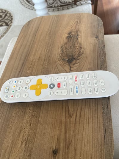 A white remote control rests on a wooden table. The remote has many buttons, including numbers 0-9, Netflix and Film Dizi buttons, directional buttons, and a yellow OK button. The wooden table has a natural grain pattern. The background is blurry, showing a couch and a patterned rug. The scene appears to be indoors during the daytime. The visible text on the remote is in English and Turkish, including "Netflix," "Film Dizi," "menü," "OK," "geri," "Çıkış," "SES," "PR," "Tv+," "kanallar," and "yazi." The overall impression is a still life of a common household object in a domestic setting.