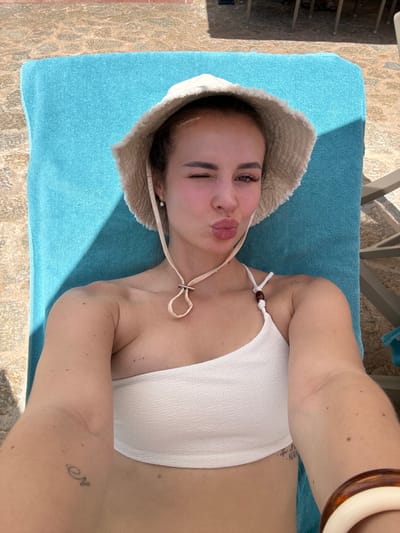 A woman lies on a blue towel on a sun lounger, wearing a white bikini top and a wide-brimmed hat. She is taking a selfie, puckering her lips and winking. The scene appears to be outdoors, with sunlight casting shadows on the towel and the textured ground. The woman has a small tattoo of a ringed planet on her left arm and another tattoo on her right arm, which appears to be text. A brown and cream-colored bracelet is visible on her right wrist. The image evokes a sense of relaxation and a holiday atmosphere, possibly during a sunny day in Es Camp de Mar, Spain.