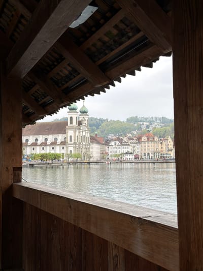 The image shows a view through a wooden structure in Lucerne, Switzerland. The perspective is framed by the dark, wooden beams and roofing of the structure, likely a bridge or covered walkway. Through the opening, a cityscape along a body of water is visible. The buildings have varied heights and architectural styles, some with light-colored facades and others with darker roofs. The River Reuss is reflecting the buildings and sky. The Baroque Jesuit Church can be seen in the foreground, with its distinctive onion-shaped domes.