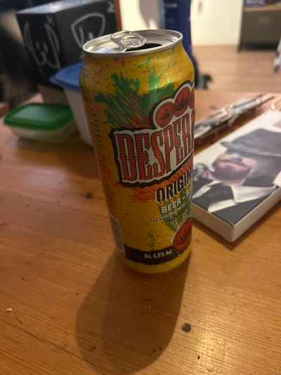 A can of "Despo" beer, labeled "Original Beer," is featured in the image. Text on the can specifies that the beer is "Flavoured with Aguardi" and contains 5.9% alcohol by volume. The can is predominantly yellow with red and green graphics. The pull tab of the can is visible, suggesting it's been opened. The can is placed on a wooden surface near an object that appears to be a book.