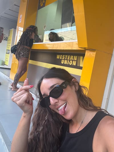 The image depicts an outdoor scene in Moo 1, Thailand, focused on a transaction point. In the immediate foreground, a woman with long, dark, wavy hair, wearing black Prada sunglasses and a black tank top, smiles broadly with her tongue slightly extended. She makes a small heart gesture with her right hand, which has red painted fingernails. A silver chain necklace is visible.

In the midground, a prominent yellow structure functions as a Western Union money transfer booth, displaying "WESTERN UNION" in black lettering. At the exterior counter of this booth, a second individual with long dreadlocks, wearing a black crop top and leopard print shorts, leans forward, seemingly interacting with a third person visible behind the glass counter inside the booth. The individual with dreadlocks has visible tattoos on their left arm and lower left leg and wears flip-flops. To the left of the main booth, another yellow sign with Thai text, including "Western Union," "Send Receive Money," and "Money Exchange," is present. The ground is a light-colored, possibly concrete or tiled surface. The scene is illuminated by bright daylight.