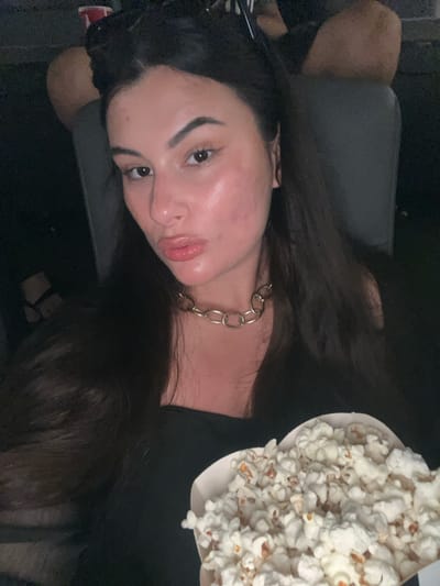 A young woman is sitting in what appears to be a dark environment. She is wearing a black shirt and a gold chain necklace. A container of popcorn is visible to her right. The image does not provide any information about the location in Kiryat Motzkin, Israel.