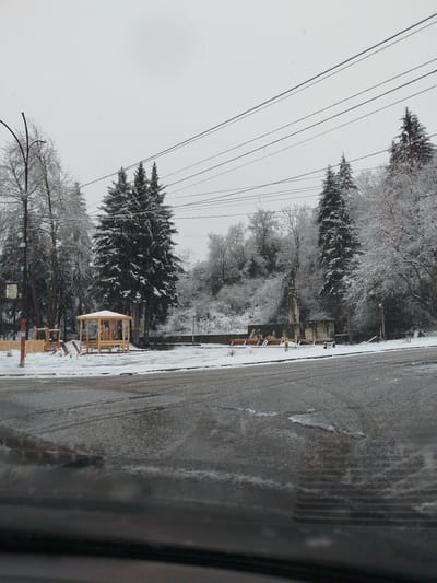 The image shows a winter scene in Klenovik, Bulgaria. It is snowing, and the trees and surrounding landscape are covered in a layer of snow. In the foreground, there is a wet asphalt road. Across the road, there is a wooden gazebo and a small play area. Further back, there are snow-covered trees, and a monument or sculpture can be seen. Overhead, there are power lines stretching across the sky. The image appears to be taken from inside a vehicle, looking out through the windshield.