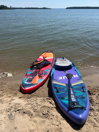 Two stand-up paddleboards are resting on a sandy shore next to a body of water. The paddleboard on the left is red with an orange and white stripe on its upper portion, featuring a colorful graphic of an astronaut and planets. An orange paddle and a black life vest are placed on this board. The paddleboard on the right is purple with a blue and yellow geometric pattern, and the word "RAVE" is visible in large white letters. A black paddle is placed on this board, and a gray bag is secured to its front. The water is calm with gentle ripples. In the background, a treeline is visible on a distant shore, and the sky is clear and blue. The image appears to have been taken during daylight hours. The scene depicts a recreational setting, likely a lake or a large river, suitable for water sports. The location is not explicitly identified as Borovoye, Russia.
