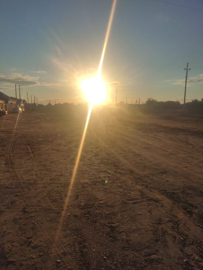 This is an outdoor, ground-level, eye-level, wide shot of a dirt road in a semi-arid environment at sunrise or sunset. The sun is low in the sky and very bright, creating a strong lens flare that stretches diagonally across the frame from the top center to the bottom right. The sky is a gradient of light blue at the top, fading to a yellowish-orange near the horizon. There are a few scattered, wispy clouds.
The foreground is a dusty, dirt road with visible tire tracks from vehicles. The ground is uneven and textured with small pebbles and debris. In the midground and background, there is a settlement with low-rise buildings, some of which appear to be residential or commercial structures with wooden siding. Several utility poles with power lines are visible, as are some sparse trees and bushes. The overall impression is of a rural or semi-urban area. The bright sun and the dry, dusty ground suggest a location that experiences a continental climate. This scene is consistent with the environment in the city of Aral, Kazakhstan, which is known for its dry climate and proximity to the former Aral Sea.