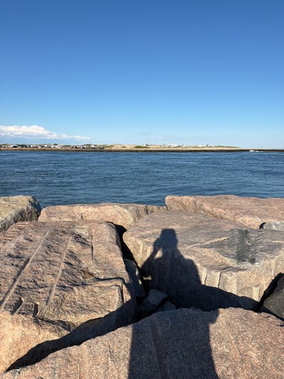 A shadow of a person holding a phone to take a picture is cast on large, rough-hewn granite rocks in the foreground. The rocks are of various sizes and shapes, some appearing to be part of a jetty or breakwater. Beyond the rocks, a body of calm, blue water stretches towards a distant shore. On the far shore, a line of sand dunes is visible, topped with vegetation and a row of recreational vehicles or campers. A clear, bright blue sky with a few wispy white clouds dominates the upper portion of the image. The scene appears to be set in a coastal area, likely Hampton Bays, United States of America, given the presence of a jetty, ocean, and beachside camping.