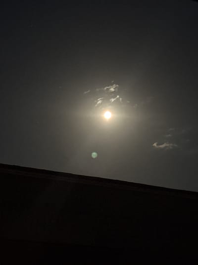 The image shows a night sky with a bright moon, partially obscured by thin clouds. Below the moon, there is a faint green light, possibly a lens flare or atmospheric phenomenon. In the foreground, there is a dark, silhouetted structure, likely a rooftop or building edge, which spans the bottom of the frame. The overall scene is dark, with the moon being the primary light source. The image was taken in Dragoman, Bulgaria.