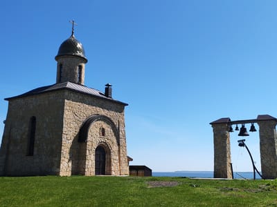 The image shows a stone chapel with a dome topped with a cross, located in Икское Устье, Russia. Next to the chapel, there is a structure with hanging bells. The chapel and bell structure are situated on a grassy area overlooking a body of water. The sky is clear and blue. There are no people visible in the image. A small shed is visible in the background.