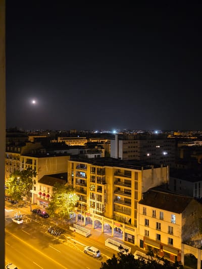 This image is a night view of a city street in Montrouge, France, taken from a high vantage point. The sky is dark with a bright full moon visible in the upper left quadrant. The foreground shows a street illuminated by streetlights, with several parked cars and motorcycles. A white car is in motion on the right side of the street, and another white car is in motion on the left side, appearing to be a police car due to its lights. Buildings line the street, with some windows lit up, indicating indoor activity. The buildings are of various heights and architectural styles, suggesting a mix of residential and commercial properties. The overall scene depicts a quiet urban night with signs of life in the illuminated windows and moving vehicles.