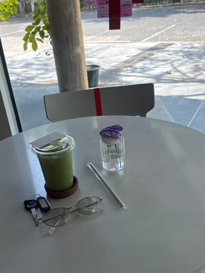 A white table displays a green beverage with a plastic lid and a white card inserted, along with a straw, a glass cylinder holding a purple flower, eyeglasses, and keys. The table is positioned near a window. A gray chair with a red stripe is visible. Outside the window, green foliage and a glimpse of a building or structure can be observed.