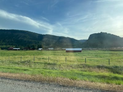 This image depicts a rural landscape in Swan Valley, United States of America. A vast, green pasture stretches across the foreground and midground, dotted with a few scattered wooden fence posts and wires. Several farm buildings are visible in the distance, including a red barn with a white roof and other structures of varying sizes and colors. Beyond the fields, lush, tree-covered mountains rise up, their slopes casting shadows in the late afternoon sun. The sky above is a mixture of blue and white clouds, with rays of sunlight breaking through. The road in the immediate foreground is gravel, with dry grass along its edge. There are no people or animals visible in the scene.