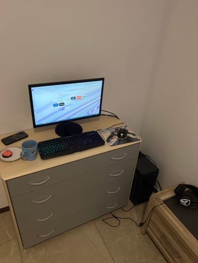 The scene depicts an indoor corner workstation within a building in Wegnez, Belgium. A light-colored, five-drawer dresser with gray drawer fronts and clear curved handles serves as a desk. On its top surface, a black computer monitor, a black RGB-backlit keyboard, and a black gaming mouse on a character-themed mousepad are arranged. The monitor displays a web browser interface with multiple shortcut icons and a purple-blue abstract background. To the left of the keyboard are a black smartphone, a light blue mug with a cartoon character design, and a small white plate holding a single cupcake with pink frosting and sprinkles. A black computer tower with an illuminated blue light is situated on the tiled floor to the right of the dresser. Adjacent to the computer tower on the right, a second piece of light wood furniture with a dark top holds a pair of black over-ear headphones. Black cables are visible connecting the electronic components and extending across the tiled floor. The walls are plain and light-colored. No individuals are present.
