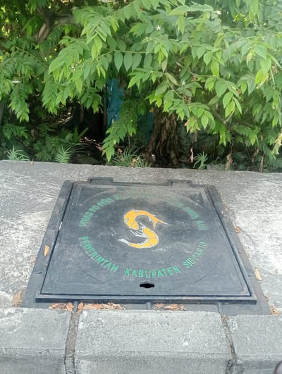 The image shows a manhole cover set into a concrete surface, likely a sidewalk or road. The manhole cover is black with green lettering and a yellow and orange symbol in the center. The lettering appears to be in Indonesian and reads "DINAS PU BINA MARGA DAN SUMBER DAYA AIR" and "PEMERINTAH KABUPATEN SIDORJO", with the year "2022" also visible. The symbol in the center is a stylized image of a serpent or dragon. Behind the manhole cover, there is dense green foliage from trees and other plants, suggesting an outdoor environment. The overall scene is mundane and depicts a piece of urban infrastructure. There are no people visible in the image. The location is identified as Sawotratap, Indonesia.