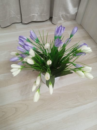 A white vase filled with artificial flowers sits on a light-colored wooden floor. The bouquet contains numerous white and lavender tulip-shaped buds, interspersed with green artificial grass. The flowers are arranged in a cluster, with some buds pointing upwards and others angled outwards. In the background, there are light beige curtains that appear to be made of a textured fabric, and a white wall is visible on the right side of the frame. The lighting suggests it is daytime, and the scene appears to be indoors, likely in a home. There are no people visible in the image, and no text is present. The overall impression is one of a decorative arrangement meant to brighten a living space.