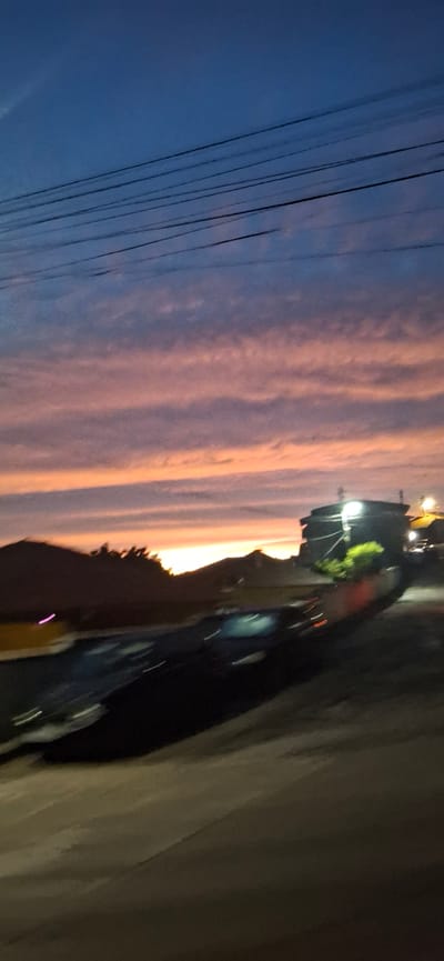 The image depicts a dramatic sunset scene with vibrant colors painting the sky over a suburban or semi-rural landscape. The sky transitions from a deep blue at the top to hues of pink, orange, and yellow near the horizon, with layered clouds catching the fading sunlight. Several dark power lines crisscross the upper portion of the frame, creating a graphic element against the colorful sky.
In the middle ground, silhouetted against the horizon, are the outlines of hills or mountains, with some vegetation visible. To the right, a building with illuminated lights stands prominently. The lower half of the image is dominated by a blurred, dark foreground, suggesting movement, possibly a vehicle or a person in motion. The motion blur makes it difficult to discern specific details of the ground or any objects within it, but it creates a dynamic, almost impressionistic feel to the scene.
The overall impression is one of a peaceful yet dynamic evening, with the natural beauty of the sunset contrasting with the man-made elements of power lines and structures. There is no indication in the image of the specific city of Valkosel, Bulgaria, or any specific details about people, actions, or interactions beyond the implied motion.