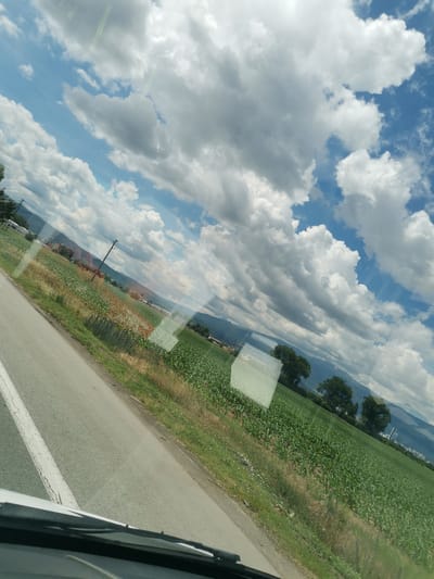A tilted, wide shot captures a view from inside a vehicle, looking out at a rural landscape under a cloudy sky. A paved road with a white line runs diagonally from the bottom left to the center left. To the right of the road, a field of green crops, possibly corn, stretches into the distance. Further back, another field with drier, lighter colored vegetation is visible. In the background, rolling hills and mountains are partially obscured by trees and more clouds. A utility pole with wires stands in the middle ground. The image appears to be taken on a sunny day with significant cloud cover, casting shadows and creating a dramatic sky. The location is identified as Krivina, Bulgaria.