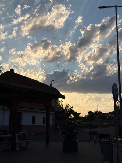 The image depicts a street scene in Arcahueja, Spain, during the late afternoon or early evening. The sky is filled with dramatic, illuminated clouds, with rays of sunlight breaking through. In the foreground, a dark silhouette of a wooden gazebo with a tiled roof stands to the left, partially obscuring a white building with a pink stripe. A stone table and benches are visible beneath the gazebo, also in shadow. To the right of the gazebo, a street lamp with a dark fixture on a tall metal pole dominates the scene. A blue circular road sign with a white arrow is attached to the pole. Further back, dark trees and houses with arched windows are visible against the bright sky. The ground in the foreground is paved with dark grey bricks. Several dark chairs and a trash can are also present on the paved area. The overall impression is one of a quiet, residential street bathed in the dramatic light of a setting sun.