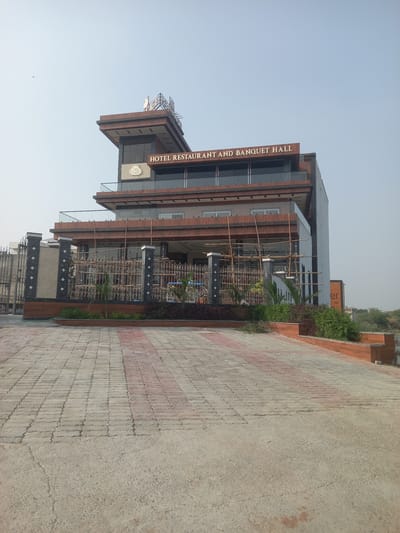 The image captures the facade of the "Hotel Restaurant and Banquet Hall" in Orai, India. The building is multi-storied with a mix of glass and brickwork. Scaffolding is present at the entrance, suggesting ongoing construction or maintenance. A patterned brick walkway leads up to the building. The sky is clear and blue. Landscaping elements, including plants, are visible near the building's base.