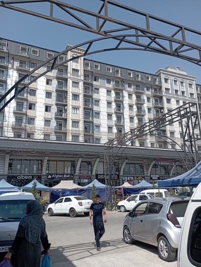 This media file captures a street scene in Termez, Uzbekistan. The foreground is dominated by a large, dark metal structure, possibly part of an awning or scaffolding, arching over the frame from the left. To the right, a multi-story building with a light-colored facade features numerous windows and balconies. The building appears to be a commercial establishment, with several signs visible at ground level. Text on these signs includes "Afghan Mall," "albem MEN'S WEAR," "SUMAYYA," and "MUSAN BOBO MARKET GOLD STAR." The sky above is a clear, bright blue, suggesting a daytime setting.
In the middle ground, several vehicles are parked along the street. A silver car is prominent, and behind it, a white car is visible. A few blue and white awnings or tents are set up along the storefronts, providing shade.
Two people are present in the scene. A man in a dark t-shirt and pants is walking or posing on the pavement, his body at an angle to the camera. Further down, a person wearing a headscarf is visible, holding a blue plastic bag. The overall atmosphere appears to be that of a busy market or shopping area on a sunny day. The perspective is slightly tilted, giving the image a dynamic feel.