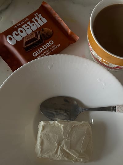 The image shows a flat-lay of a snack and drink arrangement. There is a brown chocolate bar labeled "ОСОБЫЙ" and "QUADRO" in Russian. Next to it is a white plate with a metal spoon and a block of white cream cheese. A cup with a brown liquid, likely coffee or tea, is positioned at the top left corner. The scene appears to be set on a light-colored countertop. Location: Черёмушки, Russia is speculative and cannot be confirmed from the image alone.