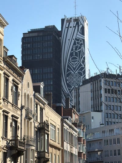 This forensic description details an urban landscape in Ixelles - Elsene, Belgium, focused on the IT Tower at Avenue Louise. The image, captured in clear daylight under a bright blue sky, features a multi-layered architectural composition.

In the immediate foreground on the left and spanning the lower half of the frame, a row of historical multi-story residential buildings is visible. These structures exhibit traditional European architectural characteristics, including beige and light brown stone or stucco facades, ornate window frames, decorative balconies with wrought-iron railings, bay windows, and visible rooftop elements like chimneys and dormers. No individuals or discernible activities are present in this section.

Dominating the mid-ground and background are several prominent high-rise buildings. A tall, dark-colored, rectangular skyscraper, the IT Tower, stands centrally, its upper section displaying a "LA VILLA IN THE SKY" sign. Adjacent to and partially overlapping this dark facade, another side of the IT Tower is prominently featured, adorned with a large, vertical, black-and-white mural. This mural depicts a highly stylized, abstract, and geometric pattern that resembles a cloaked figure or totem. To the right, additional multi-story modern buildings with numerous windows contribute to the urban skyline. The upper right portion of the frame is partially obscured by bare tree branches. The scene is illuminated by strong natural light, indicating a sunny day.