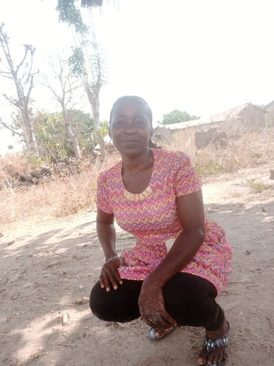 An adult female is depicted in a crouching posture on dry, light brown earth, looking directly at the camera with a slight smile. She wears a short-sleeved top with a pink, yellow, and red chevron pattern, featuring a light-colored, possibly beaded or shell, necklace along the collar. Black leggings and dark-colored flip-flops are visible on her lower body. Her hands are resting on her knees, and light-colored dust or dirt is present on her hands and lower legs. The environment, located in Gudige Sabo, Nigeria, is characterized by bright daylight. The background reveals sparse, dry, light brown vegetation and several deciduous trees, some with minimal foliage. To the right, a weathered, mud-colored structure, possibly a dwelling, with a light-colored roof is visible amidst more dry vegetation. A pile of dark rocks is present near the base of the trees on the left. Shadows are cast on the ground to the left of the subject.