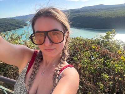 A woman is taking a selfie on a sunny day, with a large turquoise body of water and forested hills in the background. The woman has blonde hair in two braids and is wearing large, dark-rimmed sunglasses. She is also wearing a dress with a floral pattern and a cross necklace. The scene appears to be a scenic overlook, possibly a viewpoint or a park, in or near Moirans-en-Montagne, France, given the mountainous terrain and body of water. The vegetation in the foreground consists of bushes and trees.