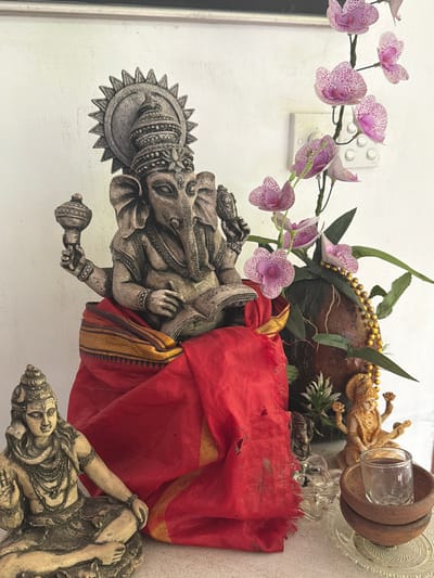 The image shows a shrine with religious figurines and flowers, likely set up in a residential or public space in Rumassala, Sri Lanka. The focal point is a large, grey stone statue of Ganesha, the elephant-headed Hindu deity, seated and holding objects in its multiple arms, with a sunburst headdress. The Ganesha statue is draped with a vibrant red cloth with gold trim. To the left, a smaller stone statue of a seated figure, possibly Shiva, is visible. To the right, pink orchids on a stem cascade over a dark, textured object, possibly a piece of bark or wood, adorned with a gold beaded garland. Behind these elements, a small golden statue of a multi-armed deity, potentially Saraswati, is positioned. In the foreground, there are small wooden bowls, a glass votive holder, and a clear glass, suggesting offerings or ritual use. The background consists of a plain, textured white wall. The overall impression is one of devotion and cultural significance.