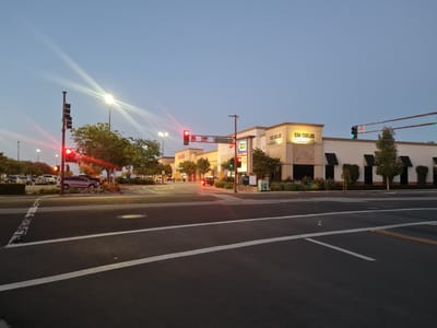 The image captures a street intersection in Redwood City, California, United States, at dusk. The sky is a clear, pale blue. In the foreground, a dark asphalt road with white lane markings is visible, indicating a multi-lane intersection. To the right, yellow lane dividers are present.

Dominating the background is a commercial building complex. A prominent sign reads "SAN CARLOS MARKETPLACE". To the left of this sign, another sign indicates "BEST BUY". The buildings are light-colored with dark awnings over some windows.

Traffic signals are visible, with a red light illuminated on a pole to the left of the frame and a green light visible on a pole further to the right. A street sign on a pole above the intersection reads "Industrial Rd". A few cars are parked in a lot behind the buildings, and trees line the street and the building's facade. One silver car is stopped at the intersection. The scene appears calm, with no pedestrians or visible activity in the immediate vicinity of the intersection.