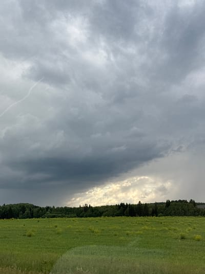 This media file depicts a natural landscape scene with dramatic cloud formations. The primary subjects are the sky and the land. The sky is filled with a variety of clouds, ranging from dark, stormy-looking masses on the left to brighter, more illuminated clouds on the right, suggesting a contrast in weather conditions or light intensity. A thin, bright line of light breaks through the darker clouds on the right side, creating a striking visual effect.

The land below consists of a dense, dark green forest that forms a distinct line across the mid-ground. Adjacent to the forest, to the right, is a vibrant green field, possibly a meadow or pasture, with scattered patches of lighter green or yellow, suggesting wildflowers or varied vegetation. The field extends towards the foreground, with a texture that indicates it is covered in grass.

The image appears to be taken outdoors, during what could be late afternoon or early evening, given the lighting. The weather appears to be transitioning, with a mix of dark, possibly heavy clouds and brighter sky visible. The location cues suggest a rural or natural setting, likely in Russia, specifically the village of Bikbarda, as indicated.

There are no visible people or objects in the frame, and no text is present. The overall mood evoked by the image is one of natural drama and perhaps a sense of anticipation due to the contrasting sky conditions. The composition, with the sharp division between the forest and the field against the dynamic sky, is visually arresting.
