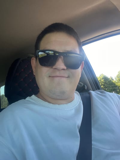 The image shows a close-up, selfie-style photograph of a man wearing sunglasses and a white sweatshirt, seated in the driver's seat of a car. The man's face is the central focus, with a slight smile. Behind him, part of the car's interior is visible, including a black seat cover with a diamond pattern and red accents, and a seatbelt. The background through the car window reveals a sunny, outdoor environment with trees and what appears to be a road with a distant sign. The context of the image suggests it was taken in Ярославка, Russia, as indicated by the prompt.