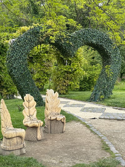 The image shows a park scene in Craiova, Romania. Three wooden, acorn-shaped benches sit beside a path. Behind the benches, a heart-shaped archway covered in green foliage stands as a decorative feature in a green area. The path is made of concrete bordered by stone and light-colored gravel. Trees and shrubbery surround the area.