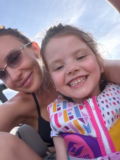 A woman with sunglasses and a child wearing a life vest are positioned close together, facing the camera. The background shows a bright sky. The woman's face is visible beneath the child's face. The life vest on the child has various colors and geometric shapes. The photo appears to be taken in an upward angle. No information is available to confirm location.
