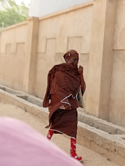 The scene is set in Gayawa, Nigeria, depicting an individual walking on a dirt path alongside a building. The person, an adult, is dressed in a brown patterned head covering and a matching draped garment with white trim. Bright red socks adorned with white patterns are worn beneath silver open-toed sandals. A patterned fabric bag is carried on the left side, and the right hand is raised towards the face. To the right of the path, a light-colored building wall featuring rectangular relief patterns extends horizontally, parallel to a concrete drainage channel. The ground consists of light brown exposed earth. Blurred green foliage and a bright sky are visible in the upper background. A soft pink, out-of-focus object occupies the lower-left foreground.