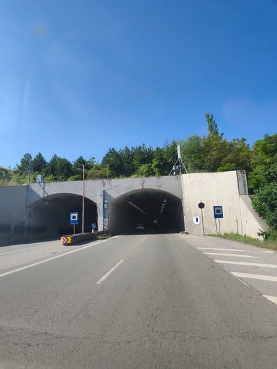 The image shows the entrance to a tunnel on a sunny day. The tunnel has two openings, side-by-side, with a concrete structure surrounding them. Road markings are visible on the asphalt, including white lines. There are traffic signs present, one indicating a tunnel entrance and another with a directional arrow. A yellow and red chevron warning sign is also visible on the left side of the road. Trees and vegetation are seen behind the tunnel entrance, and the sky is clear blue. The scene appears to be on a road leading into the tunnel.
