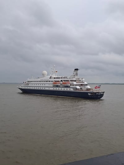 The image shows a ship, the SeaDream Yacht Club vessel, in the port of Brunsbüttel, Germany. The scene is characterized by overcast weather and a river, likely the Elbe, reflecting the dull light. The ship, with its black and white color scheme and orange lifeboats, is prominently displayed, with the Danish flag visible at the stern. The foreground includes a textured, light-colored surface.