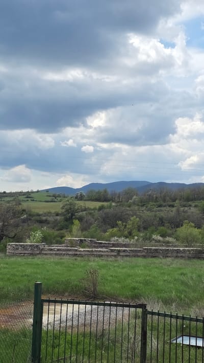 A vibrant landscape image, likely taken in Drenkovo, Bulgaria, showcases a rural mountainous scene under a partly cloudy sky. The upper portion of the frame is dominated by a bright sky featuring large, dynamic white and grey cumulus clouds, with patches of clear blue sky visible between them.
A range of forested mountains or hills stretches across the middle ground, displaying various shades of green foliage. Below the distant mountains, a valley unfolds with expansive, lush green fields. Scattered trees, some with white blossoms, are interspersed throughout the landscape, adding texture and lighter tones.
In the mid-ground on the right, ancient-looking stone or brick structures, possibly remnants of historical buildings or terraced foundations, are partially covered by vegetation. Closer to the viewer, along the right edge of the foreground, a dark green metal fence with vertical bars stands, partially obscuring a gravel path or dirt road. Faint utility lines are visible, vertically traversing the sky.
The scene is brightly illuminated, indicating daytime, likely morning or afternoon. The overall atmosphere is peaceful and serene, with no human or animal activity explicitly depicted. No visible text is present in the image.