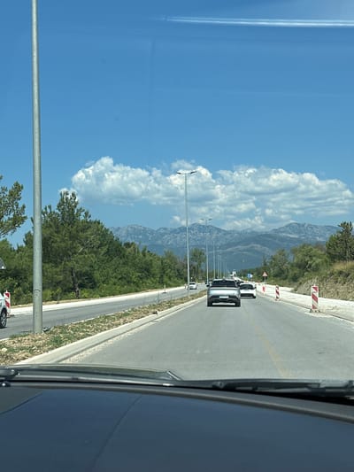 The image shows a view from inside a vehicle looking out at a road in Bratešići, Montenegro. The road is paved and has a yellow lane marker. There are several cars on the road, including a silver SUV in the foreground. To the left of the road, there is a sidewalk and a grassy area with trees. In the background, there are mountains and a blue sky with white clouds. There are also construction barriers on the right side of the road. The scene appears to be a sunny day.