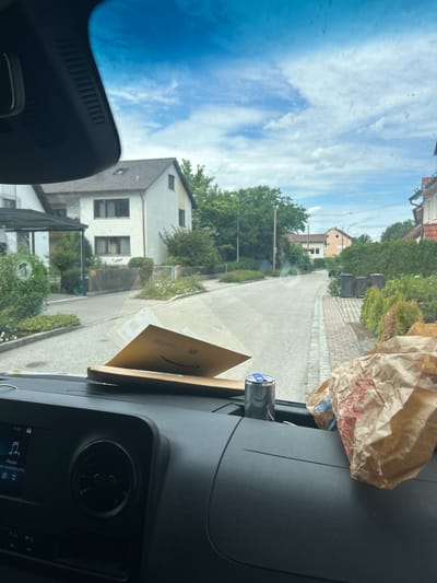 The image presents a view from inside a vehicle looking out through its windshield onto a residential street in Moosburg, Germany, under a partly cloudy sky.
In the immediate foreground, the black dashboard of the vehicle is visible. On the left side of the dashboard, an infotainment screen displays "15:39" and music playback controls, including a musical note icon and text in Cyrillic script (e.g., "Медиа"). To the right of the screen, a circular air vent is visible. Centered on the dashboard, a brown Amazon padded envelope, partially open, rests with its logo and shipping information visible. Further to the right, a silver-blue energy drink can (Red Bull) is placed in a cup holder or storage slot, and a crumpled brown paper bag, with red text partially legible, sits on the passenger side of the dashboard. The windshield itself exhibits some minor smudges and reflections.
The exterior scene reveals a two-lane asphalt road that gently curves to the right in the distance. A paved sidewalk made of light-colored pavers runs along the right side of the street.
On the left side of the street, a white, multi-story house with a brown tiled roof and dark-framed windows is prominent, featuring a carport or covered entrance area. Lush green bushes and trees surround this property. A tall wooden utility pole with a street light fixture stands on the left.
On the right side of the street, closer to the foreground, another house with a brown tiled roof and light-colored walls, featuring a distinctive yellow section, is visible. Further down the street on the right, three dark-colored trash bins are positioned near the curb, backed by a hedge and smaller green shrubs. In the background, additional residential structures with tiled roofs are discernible. The overall environment is characterized by well-maintained houses, green vegetation, and a typical suburban street layout. The sky is bright blue with scattered white clouds, indicating daytime conditions.