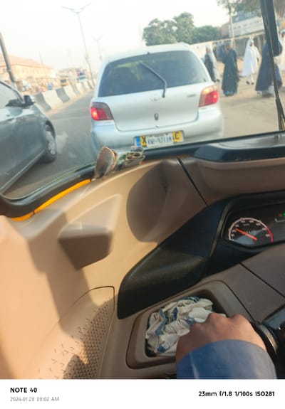 From the perspective inside a car, a silver Toyota Yaris with license plate XKW-921AM is seen in front, followed by another car on the left. Pedestrians dressed in traditional garb walk nearby. The driver's hand and dashboard are visible. The speedometer shows a speed around 40 km/h. A crumpled cloth sits in a compartment. The photo was taken with a NOTE 40 at f/1.8, 1/100s, ISO281, on January 28, 2026, at 8:02 AM.
