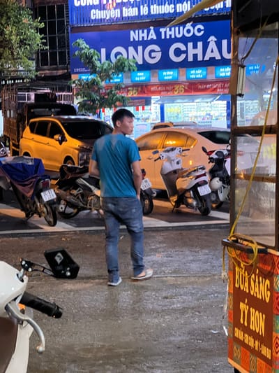 In the city of Bắc Hà, Vietnam, on a wet street at night, a person wearing a blue t-shirt and jeans stands looking to the right. In the background, a yellow car is parked alongside several motorcycles. A pharmacy sign reads "NHÀ THUỐC LONG CHÂU." To the right, a cart with an orange and brown patterned side displays text in yellow that includes "BỮA SÁNG TÝ HON". The street is reflecting the surrounding lights, indicating it has recently rained.