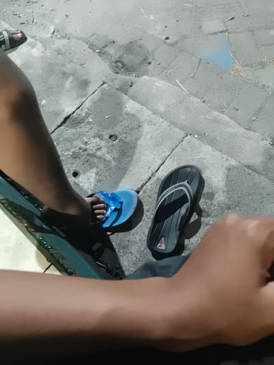 In Barengkrajan, Indonesia, an image depicts a close-up of two pairs of flip-flops on a concrete surface. One pair, a blue "anda" brand, is worn by a person whose bare foot is visible. The other pair, a dark gray flip-flop with a lighter gray stripe and a red logo, lies on the concrete nearby. The scene appears to be outdoors, with cracked concrete paving and scattered debris suggesting a public or less-maintained area. Shadows indicate it is likely evening or night. The perspective is from a low angle, possibly from someone sitting or lying down.