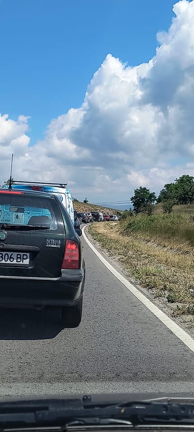 The image depicts a traffic jam on a two-lane road with a white line marking the lanes. The foreground shows the rear of a dark green car with a license plate reading "306 BP" and the word "OCTAVIA" on the trunk. To the left of this car is a white van with blue graphics. A traffic queue extends into the distance, with various vehicles visible, including cars and vans. The road is bordered by dry, grassy hills with scattered green trees. The sky above is a vibrant blue with fluffy white clouds. The scene is consistent with a summer day, likely in a rural or semi-rural area, given the natural landscape. The specific location in the city of Ponor, Bulgaria, is not discernible from the visual information provided in the image.