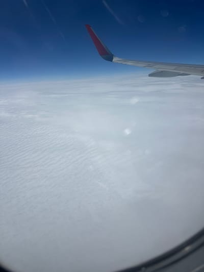 The image shows a view from an airplane window looking out at clouds and the sky. A portion of the airplane wing, with a red tip, is visible in the upper right corner. The clouds below are dense and appear to have a textured, rippled surface. The sky is a clear, deep blue. There is no information in the image to suggest a location in the city of Sim, Russia. The scene is devoid of people, specific objects, or identifiable actions or interactions.