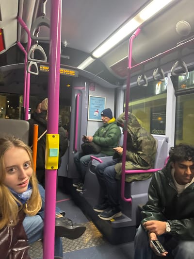 Inside a bus, several individuals are occupied. A young woman with blonde hair and a maroon jacket gazes forward. Behind her, a row of passengers are seated, including two men in green jackets and another man wearing a green leather jacket, checking his watch and using a phone. A purple bar runs across the frame, and the bus interior is lit with ceiling lights. The overall scene appears to be a candid snapshot of daily commuting.